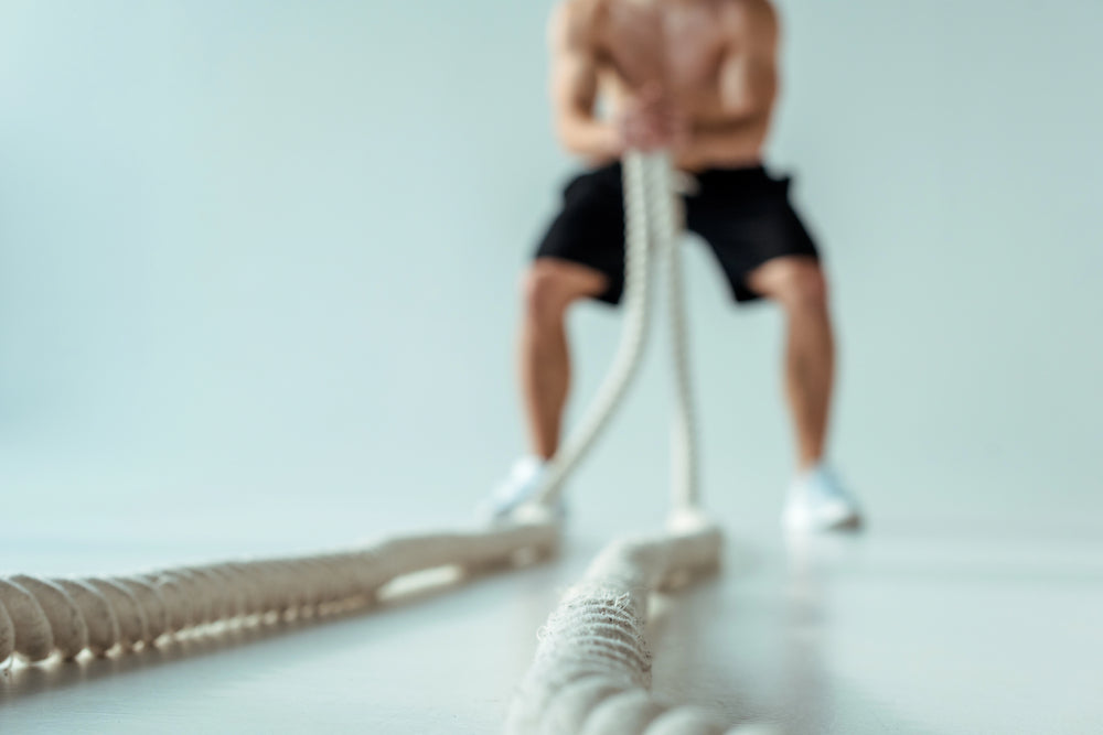 Selective focus of sexy muscular bodybuilder with bare torso exercising with battle rope on grey background